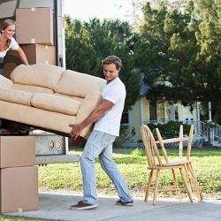 A young couple lifts a tan couch to move it in or out of the back of a moving truck.  A young woman with blond hair pulled away from her face is standing inside the truck holding the left end of the couch.  The woman wears a white top and dark blue jeans.  A young man with brown hair stands outside the truck holding the right end of the couch.  The man is wearing a white shirt and light blue jeans.  Moving boxes and sit inside and outside the truck.  Two chairs sit on a sidewalk outside the truck.  A yellow house can be seen in the background.