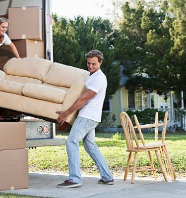 A young couple lifts a tan couch to move it in or out of the back of a moving truck.  A young woman with blond hair pulled away from her face is standing inside the truck holding the left end of the couch.  The woman wears a white top and dark blue jeans.  A young man with brown hair stands outside the truck holding the right end of the couch.  The man is wearing a white shirt and light blue jeans.  Moving boxes and sit inside and outside the truck.  Two chairs sit on a sidewalk outside the truck.  A yellow house can be seen in the background.